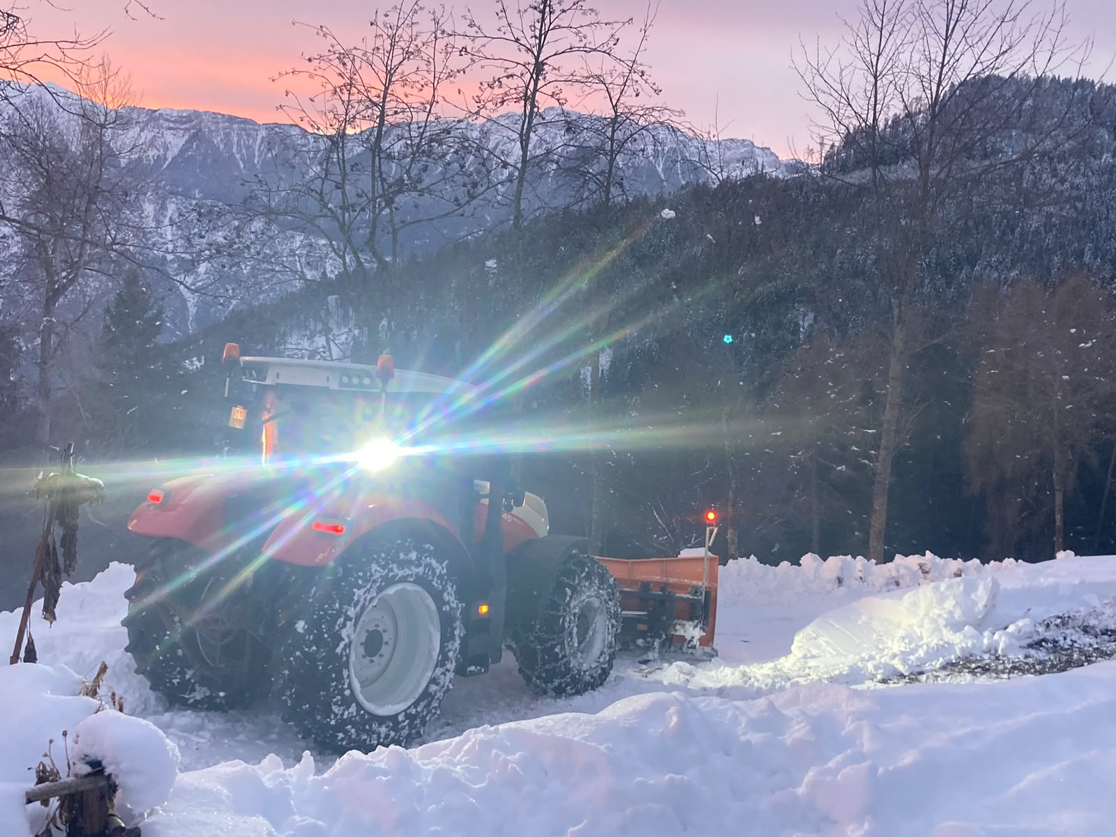 Trattore con lama spazzaneve al lavoro all'alba, tra la neve e le montagne
